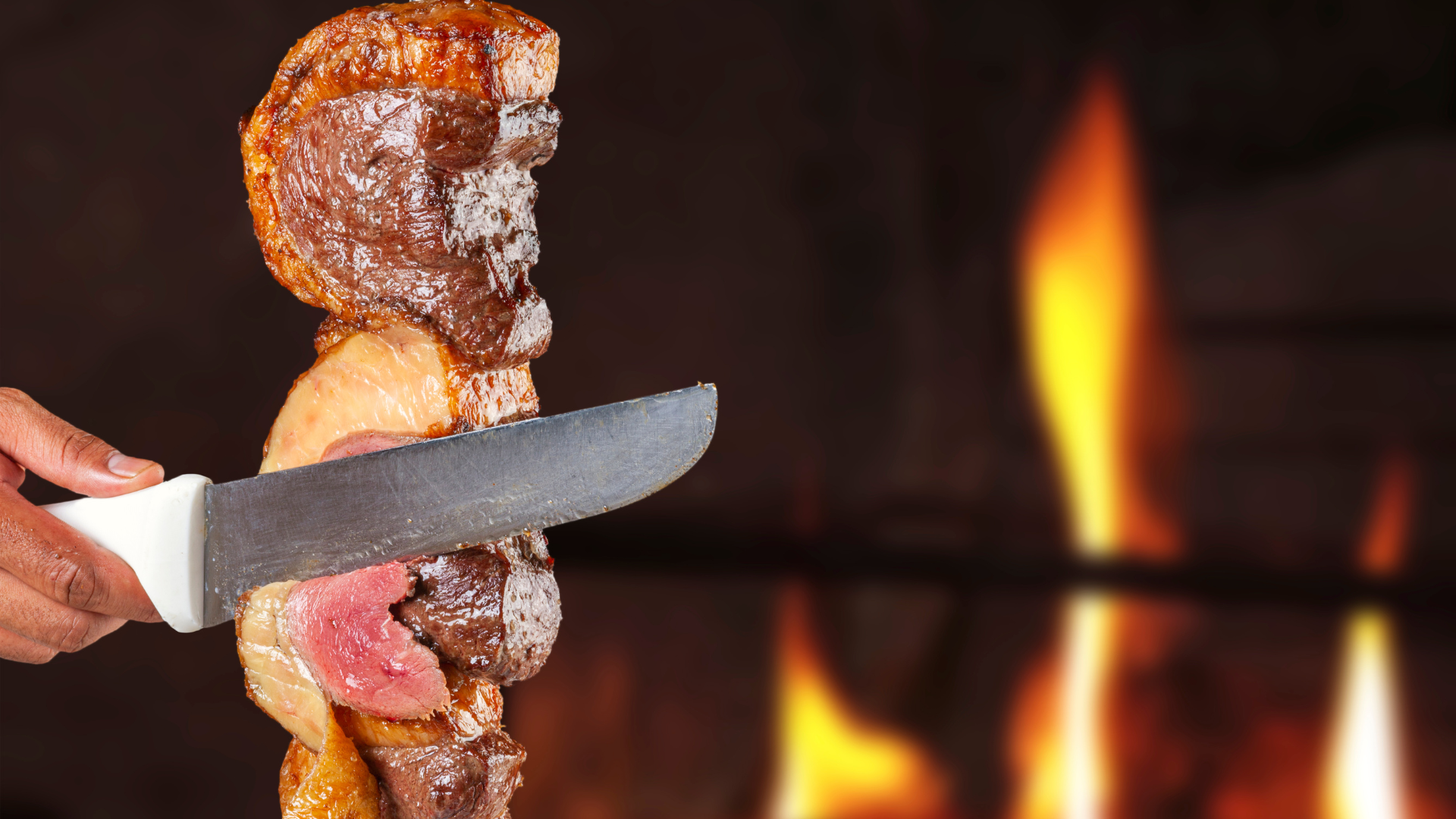 Espeto de picanha a ser cortado na mesa, com o fogo da churrasqueira ao fundo